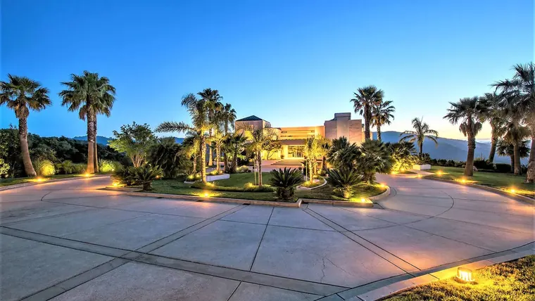 A contemporary estate at twilight, its circular motor court lined with lit palms and the hills behind it