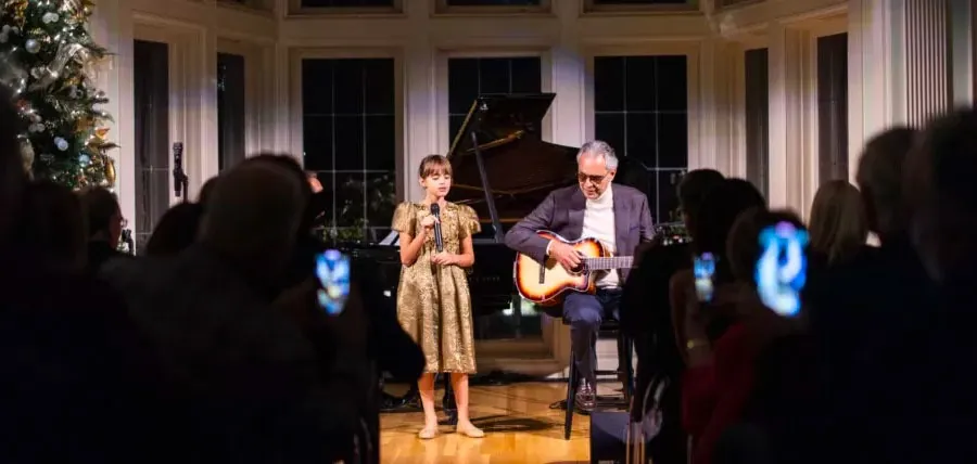 Andrea Bocelli plays guitar beside a young singer in a gold dress at an intimate candlelit concert